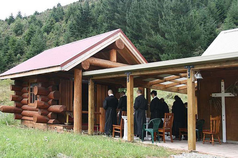 Die Kapelle des Stützpunktes der FSSR in Christchurch, Neuseeland und das daneben teilweise sichtbare Wohnhaus sind als Blockhäuser ausgeführt. Vor der Kapelle warten einige Brüder auf den Einzug zum Gebet.