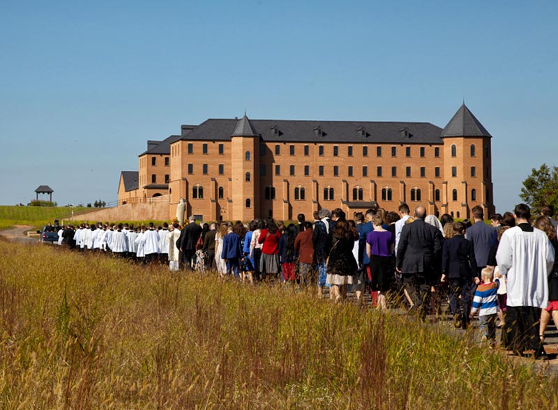 Das Thomas-Aquinas-Seminar der Piusbruderschaft in Dilwyn, Virginia. Die Anlage ist noch nicht fertig. Auf dem Platz bis zum freistehenden „Glockengestell“ links hinten soll die große Kirche entstehen.
<br>Bild: FSSPX Wie eine Festung erhebt sich der Bau des Seminars aus der Landschaft, dem eine Prozession von Wallfahrern im Vordergrund zustrebt. <br>Bild: FSSPX