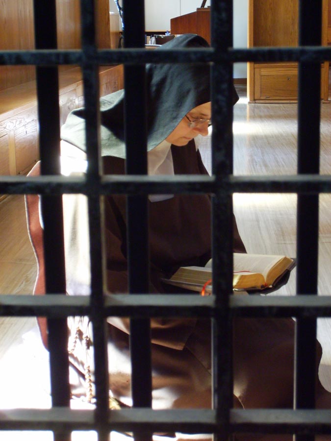 Nicht die die Nonne ist eingesperrt, sondern das Chaos der Welt soll ausgesperrt bleiben. Zum Heil der Seelen und der höheren Ehre Gottes. <br>Bild: http://carmelitesofrochester.org. Das Photo zeigt eine Nonne im traditionellen Habit der Karmeliterinnen, die hinter dem Klausurgitter sitzend das Brevier betet.