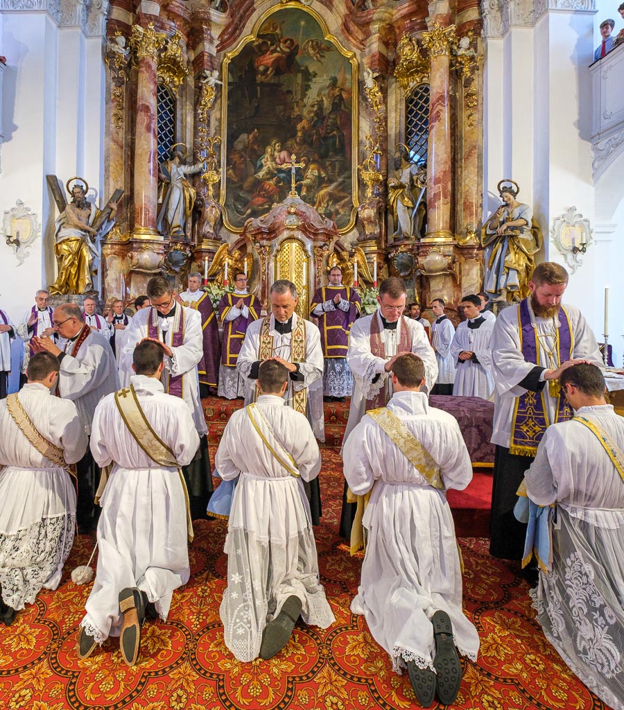 Bild: Website des Seminars Wigratzbad Die Neugeweihten knien in einer Reihe vor dem Altar, während alle anwesenden Priester ihnen ebenfalls die Hände auflegen.