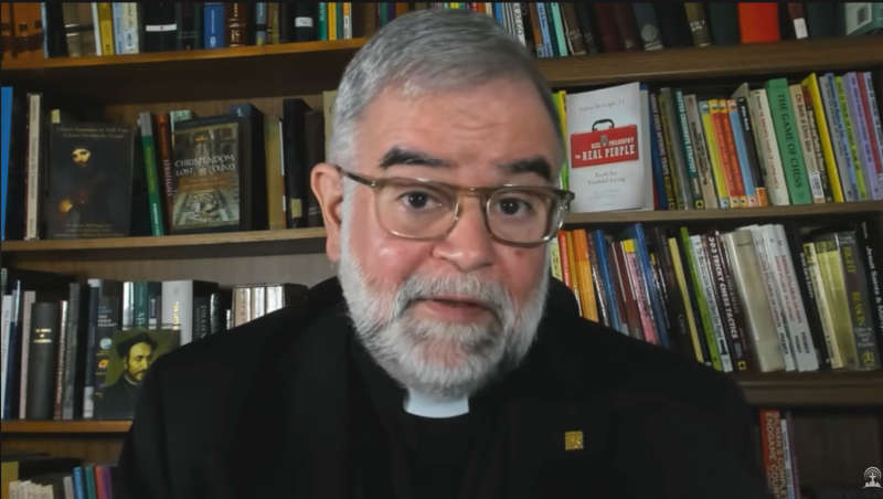 Fr. McTeigue bei der Aufnahme seines Podcasts vor dem Hointergrund seiner Bücherwand - mit zahlreichen eigenen Titeln