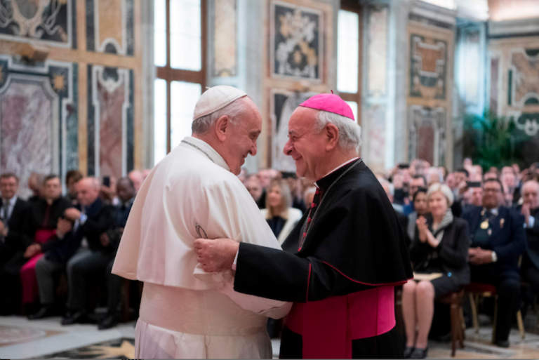Herzliche Umarmung bei einer Begrüßung von Papst Franziskus und Erzbischof Paglia vor dem Hintergrund einer mit Würdenträgern besetzten Aula.